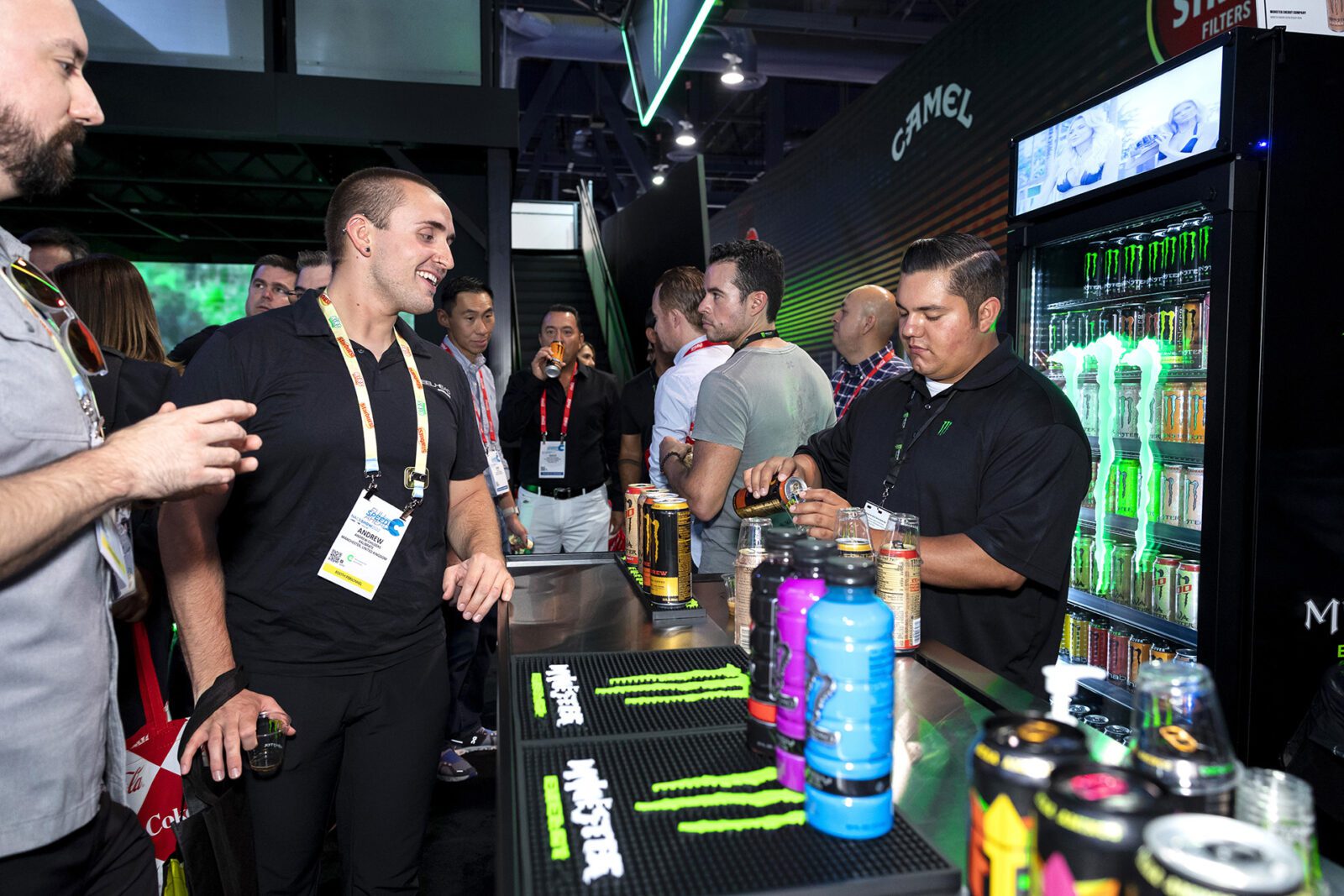 Monster ED - 5 People gather at a Monster Energy booth where a staff member prepares drinks; energy drink cans are displayed in a fridge and on the counter.