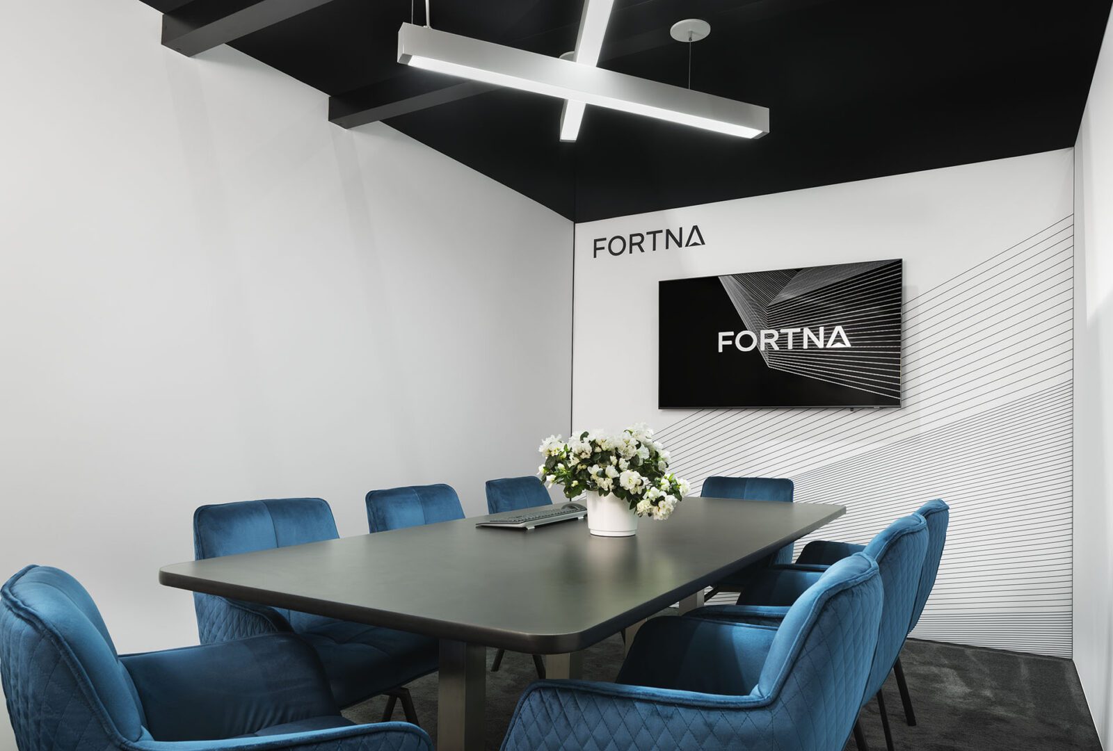 A modern conference room with a black table, blue quilted chairs, a floral centerpiece, and a wall-mounted screen displaying "FORTNA"—an award-winning introduction of the NEW Fortna brand to the world at ProMat 2023!.
