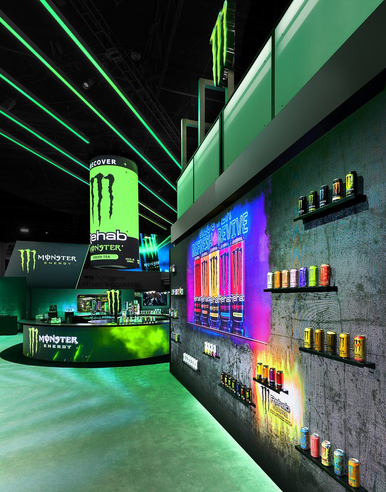 A brightly lit Monster Energy booth displays various energy drink cans on shelves and a vibrant wall, with a counter and large Monster logos throughout the space.
