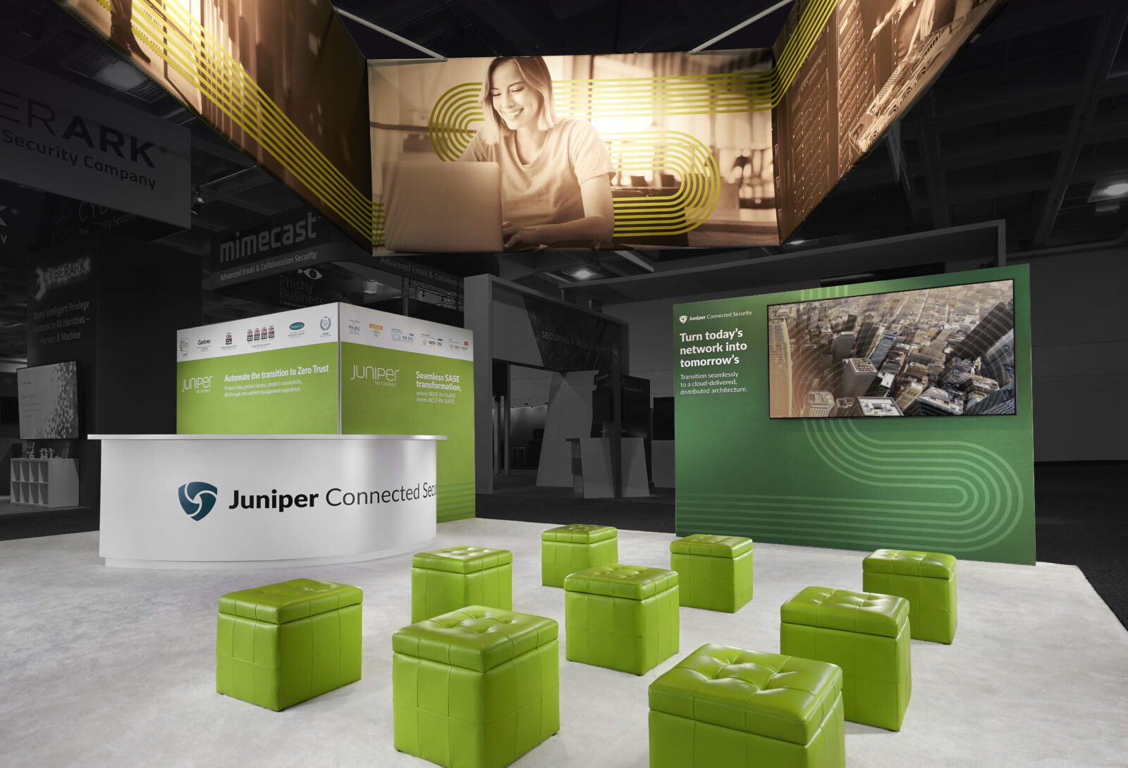 2023_RSA_Juniper_03 Trade show booth with green cube seats, Juniper branding, digital screens, and large overhead banners featuring a woman and cityscape—an engaging trade show marketing setup designed to capture attention.