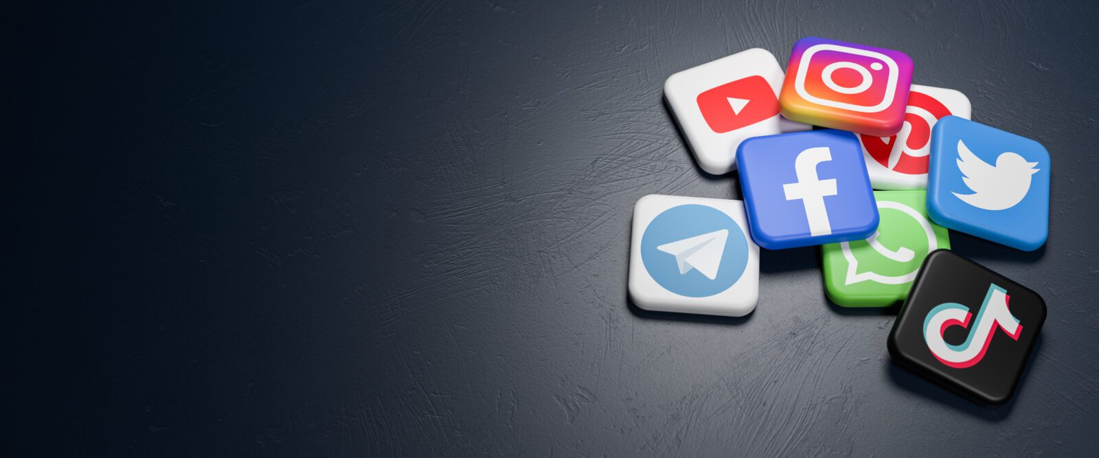 Logos of the major social media and messaging sites (Youtube, Instagram, Pinterest, Twitter, Facebook, Whatsapp, Telegram, Tiktok) on a heap on a dark table. Copy space - Web banner size