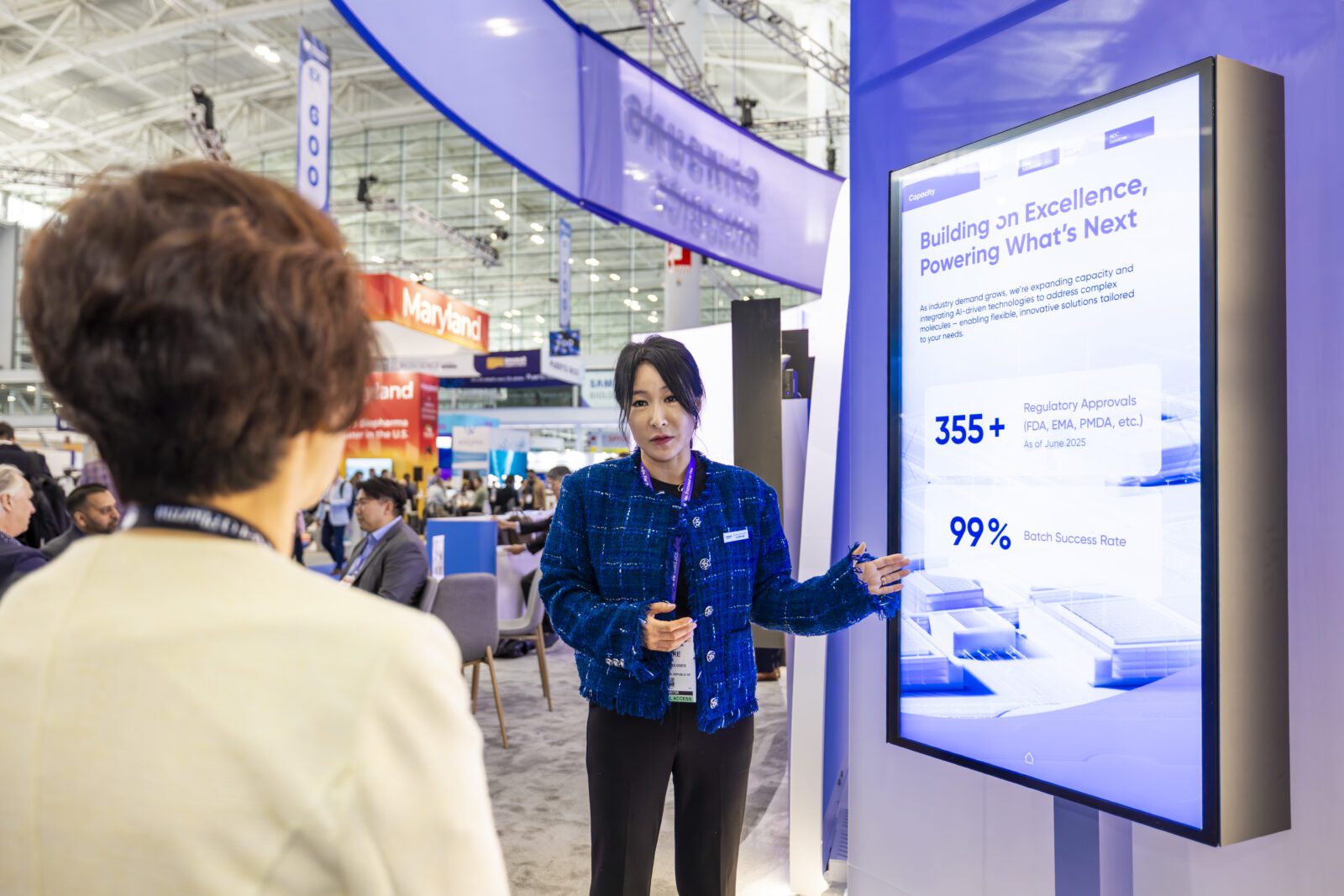 A woman in business attire presents data on a large digital screen to another person at a professional conference or exhibition.