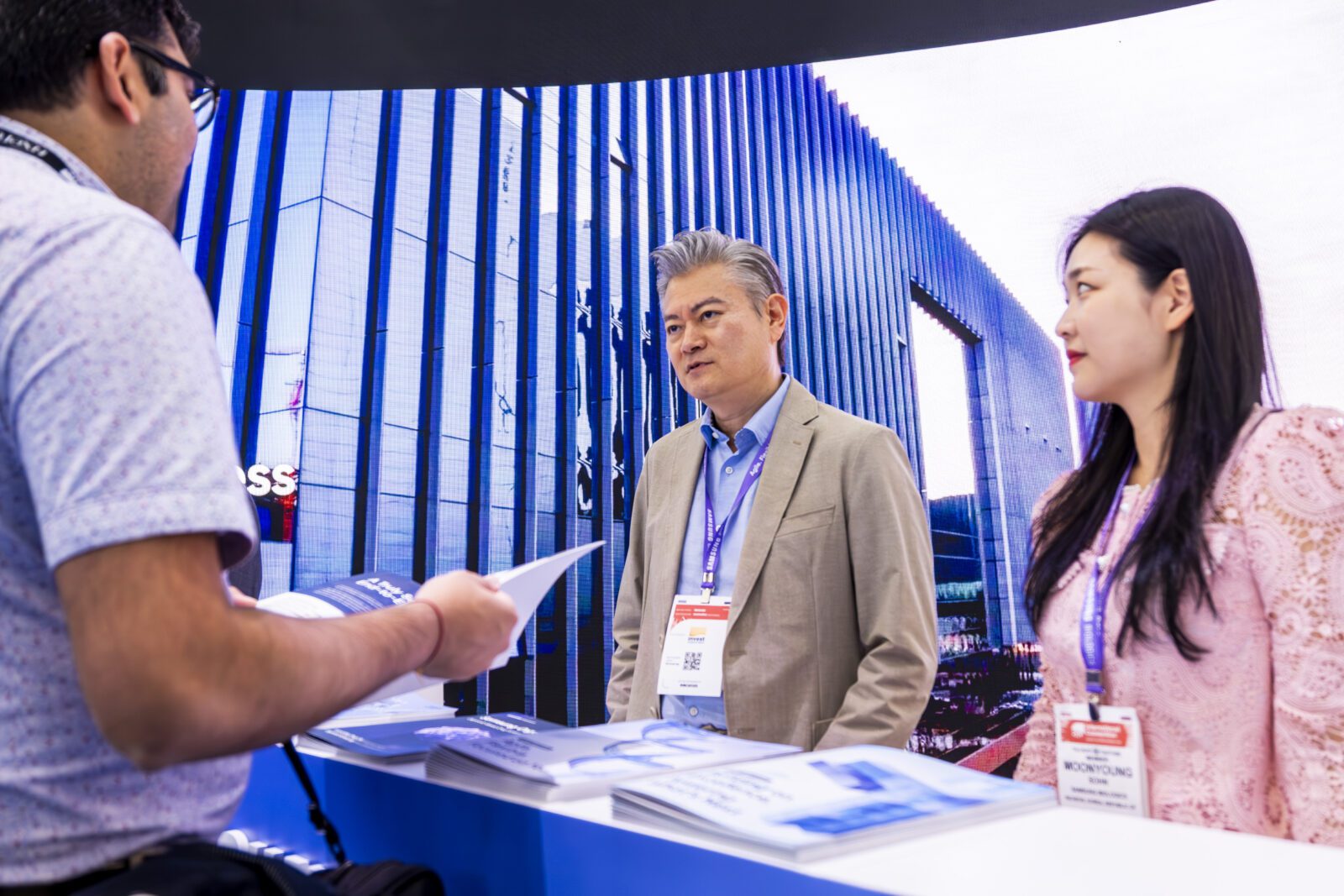 Two people stand behind a counter at a booth, speaking to a visitor holding brochures in front of a display showing a modern building exterior.