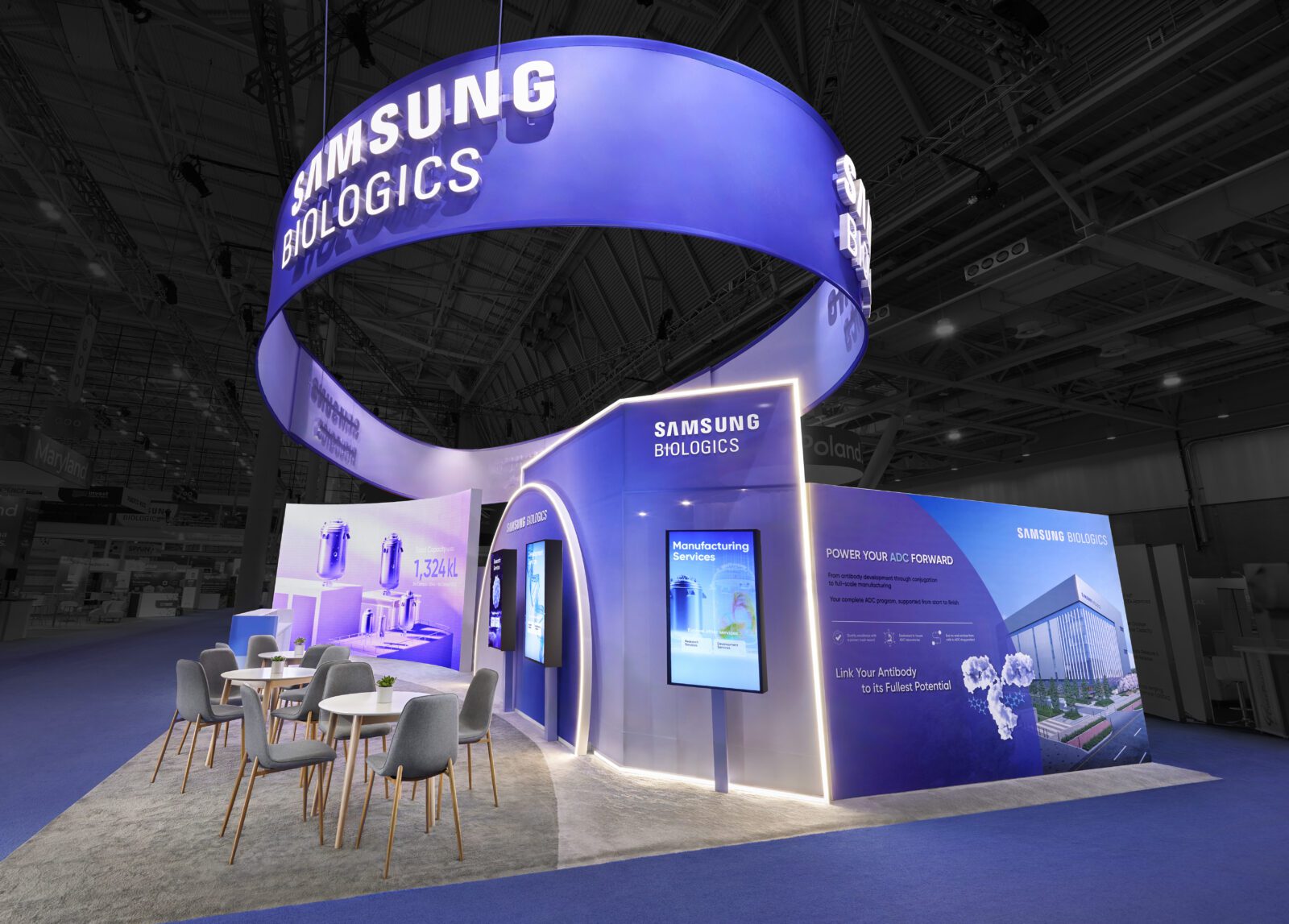 Exhibition booth for Samsung Biologics with digital displays, informational graphics, seating area, and large illuminated signage overhead.
