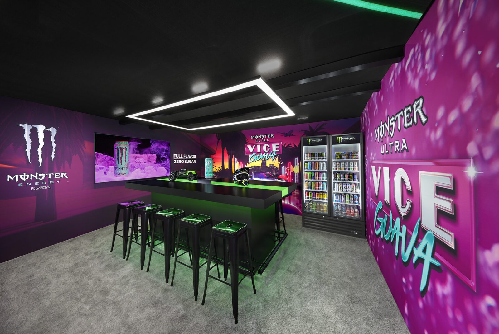 A modern room with bright pink Monster Energy branding, a bar counter with stools, beverage fridges, and neon lighting, promoting Monster Ultra Vice Guava flavor.