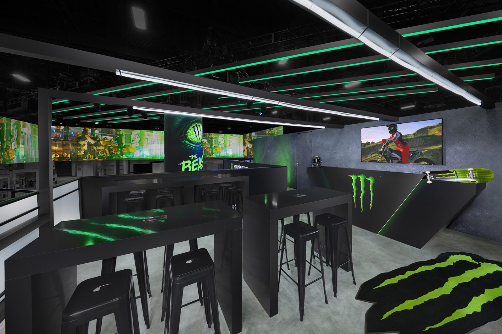 A modern lounge area with black tables and stools, green LED lights, and Monster Energy branding on walls, counters, and a rug. A TV screen displays a motocross scene.