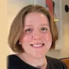 A person with short light brown hair and a black top smiles at the camera in an indoor setting with shelves and wall decor in the background.