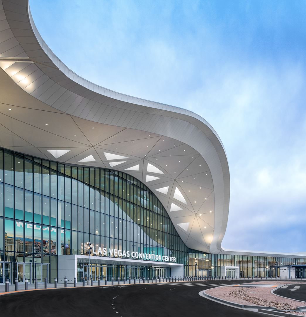 Exterior view of the Las Vegas Convention Center featuring a modern, curved roof structure, large glass windows, and an empty driveway in front.