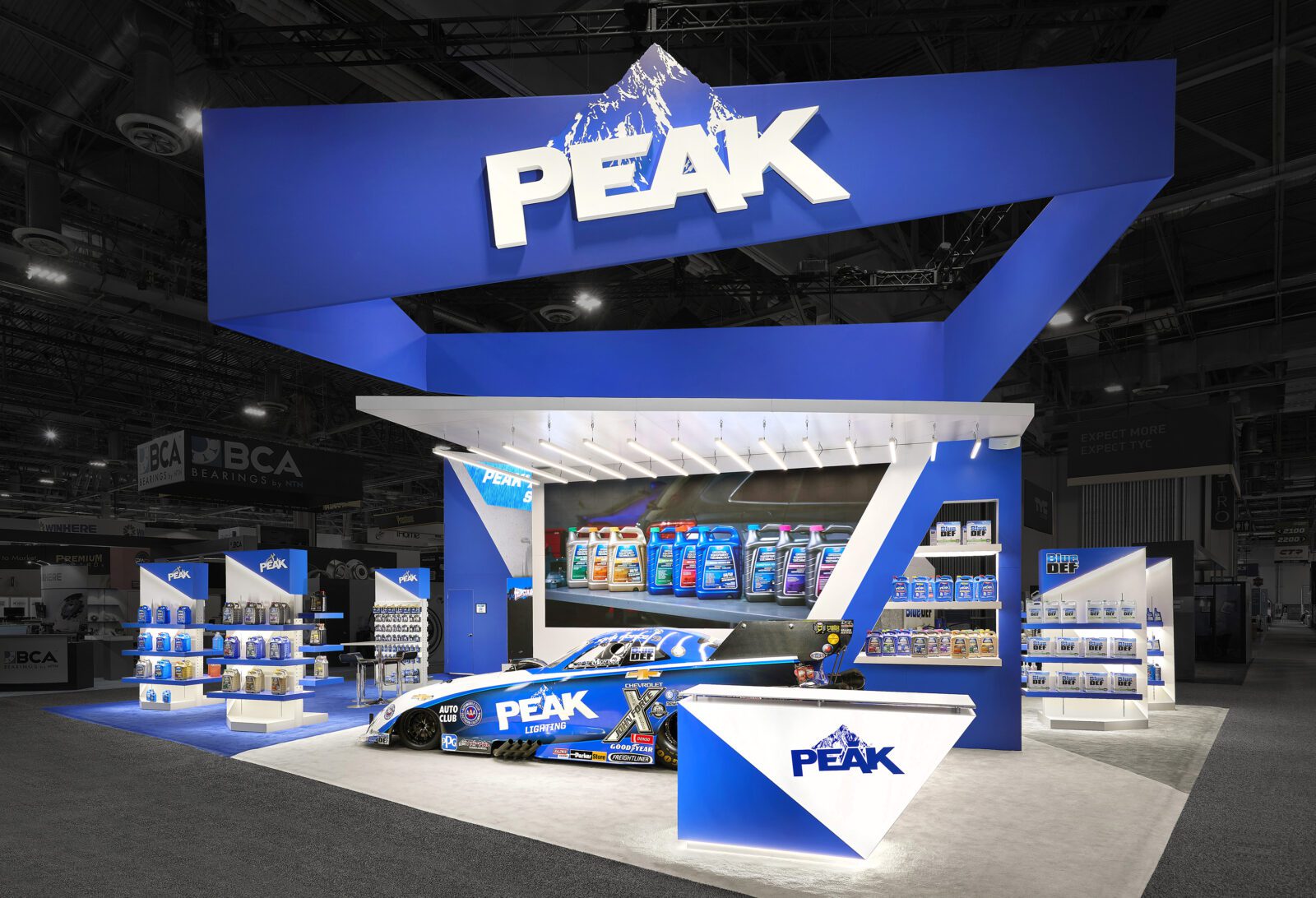 Trade show booth for PEAK, featuring branded displays, automotive products, a race car with company logo, and a blue and white color scheme.