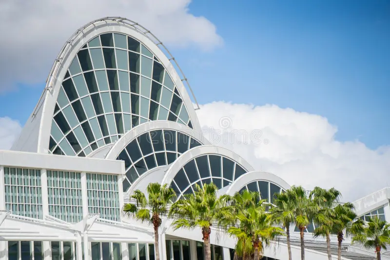 Modern building with curved glass and steel arches, set against a blue sky with white clouds and lined with palm trees in the foreground.