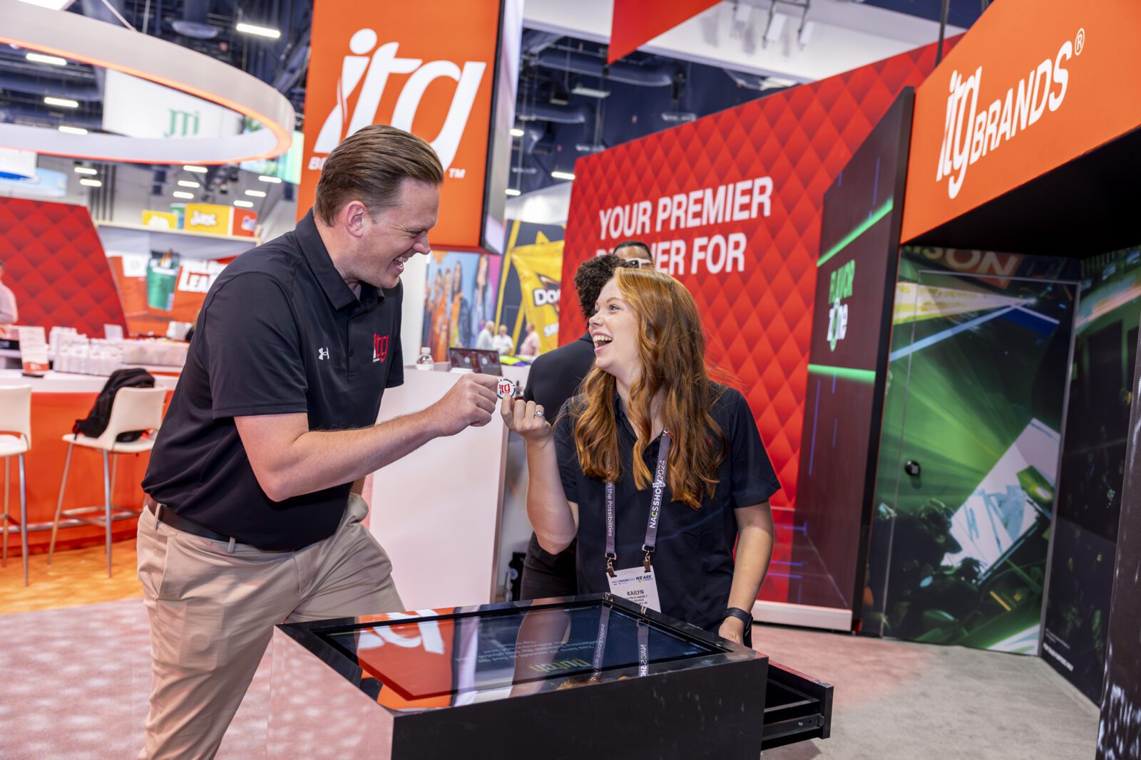 Two people in black shirts smile and fist bump at an ITG Brands trade show booth with red branding in the background.
