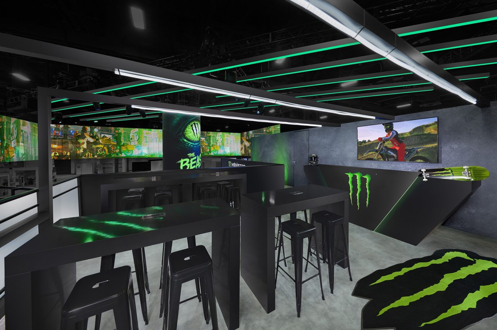 A modern gaming lounge, inspired by cutting-edge trade show design, features black tables, stools, green Monster Energy branding, neon lighting, and gaming screens on the walls.